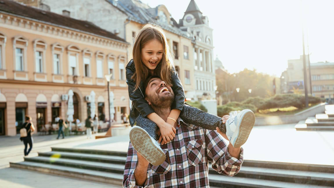 A father carries his daughter on his shoulders through a town square in a European city.